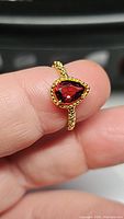 Close-up photo of pear-shaped garnet ring held between fingers showing red orange pink gemstone with twisted gold-plated sterling silver band.