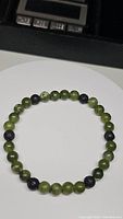 Top-down photo of natural green jade and black lava stone bead bracelet on a white surface, showing round beads and elasticity.