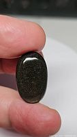 Natural gold sheen obsidian oval cabochon gemstone being held between fingers, showing dark base color with fine golden flashes and reflective surface.