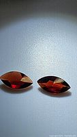 Close-up image of two marquis cut, deep reddish-brown natural almandine garnets placed side by side on white surface.