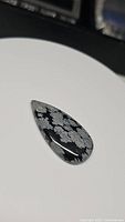 Close-up image of pear-shaped natural snowflake obsidian cabochon with distinct white snowflake patterns on a black background.
