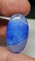 Close-up photo of natural blue striped agate oval cabochon held between fingers, showing blue and white banded patterns and polished surface.