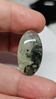Close-up photo of oval cabochon rutilated prehnite held between fingers showing mossy green to black needle-like inclusions against pale green translucent background.