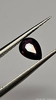 Pear-shaped deep red purple pink sapphire held between tweezers showing facets and color