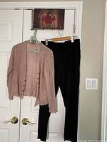 Black casual pants and light pink cardigan hanging on hangers against a white door, showing full front view of both items.