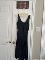 Front full-length view of the black sleeveless dress hanging on a hanger against a door, showing the full skirt and scoop neckline.