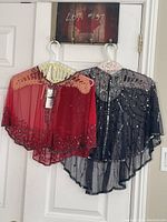 Two sheer embellished shawls on hangers against a white door, showing red and blue colors with bead and sequin decoration.
