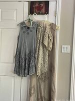Wide shot of the three items hanging on hooks: short blue/gray dress, taupe lace maxi dress, and taupe sparkly sweater.