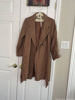 Front view of camel coloured medium coat hanging on a hanger showing double-breasted front, wide lapels, and belt end at bottom.