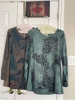 Two Casa Donna long sleeve tops hanging side by side on door hooks, showing front view with detailed feather and animal prints, one teal green and one brown.
