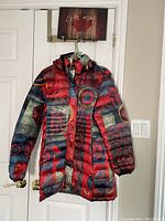 Front view of colorful quilted winter jacket hanging on a door, showing hood, zipper, and patchwork design in red, black, green colors with circular and abstract patterns.