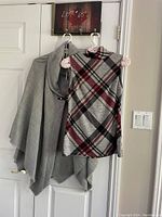 Grey poncho with cowl neck and buckle clasp hanging on a door alongside the plaid sleeveless top.