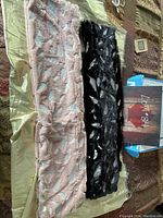 Two infinity scarves, one light pink and one black, laid flat side by side showing the metallic feather design and texture.