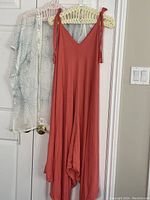 Photo showing orange sleeveless jumpsuit hanging in front of a sheer white jacket with turquoise details placed on a door.