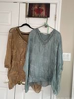 Both taupe and blue flowy tops hanging on hangers on a door, showing front view and details.