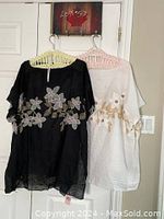 Photo showing two pullover shawls hanging on hangers, one black with floral embroidery and one white with floral embroidery.