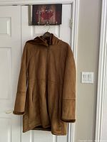 Front view of long camel color suede coat with zipper and hood, showing structured seams and two side pockets.