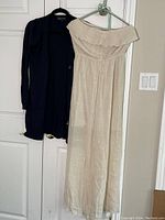 White strapless summer dress hanging next to black long sweater with pockets, both displayed on hangers against white door.