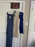 Three sleeveless garments hanging side-by-side on hangers on a door: beige long vest, royal blue top, and blue maxi sundress.