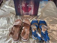 Two pairs of wedge sandals on a shiny fabric background, cream EOS pair and blue Fly pair, with a decorative maple leaf board behind.