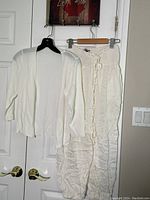 Front view of the white cardigan sweater and the white drawstring pants hanging on a wall-mounted rack, showing the texture and fit of the fabric.