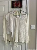 Four white garments hanging on hangers on a door, showing from back to front a long sleeve blouse, short sleeve blouse, white pull over shawl with fringe trim, and sleeveless knit tank top.