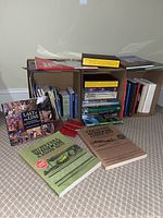 Books and magazines stacked and partially displayed on a cardboard shelf on a carpeted floor. Various titles and sizes visible including large softcover books and magazines.