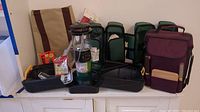 Photo showing the complete lot including Coleman propane lantern on a black base, three wine bottle totes in green and maroon colors each containing glasses and stoppers, manual kerosene pump, and bottle of Ben's Tick and Insect Repellent.