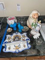 Overall view of the lot showing a ceramic lamb cookie jar, two blue rooster figurines, an espresso cup and saucer, a glass case, porcelain jar, dog figurine, wall hanging shelf and small dish on a countertop.