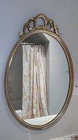 Front view of the vintage gilt oval mirror mounted on a wall with visible gold painted wood frame and ornate Victorian style top crest.