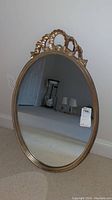 Front angled view showing oval mirror with gilt carved crest and gold painted wooden frame. Mirror reflects room interior.