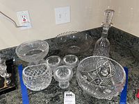 Full lot of glass items on counter including two large serving bowls, tall decanter with stopper, three small goblet-style glasses