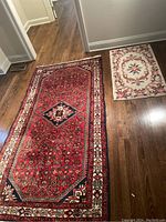 Wide view showing both rugs laid out on hardwood floor. Larger rug in red with geometric design, smaller cream floral rug side by side.
