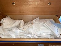 Drawer filled with assorted folded white table linens, showing various textures and layers inside a wooden drawer.