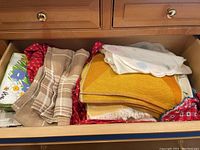Drawer packed with folded table linens including a beige plaid, red patterned fabric, yellow woven linen with fringe, white linen with pastel embroidery