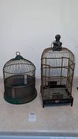 Photo showing both vintage bird cages side by side on the floor against a white wall.