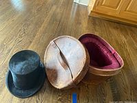 Round vintage brown leather hat storage box with handle, showing wear and burgundy padded interior lining.