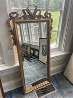 Full view of the tall rectangular wall mirror leaning against a window showing the mirror and gold painted frame with ribbon-style design on top and sides.