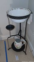 Full view of enamel wash basin seated in black wrought iron stand with matching enamel pitcher on lower shelf.