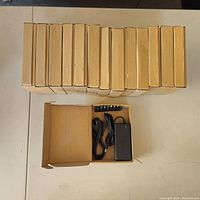 14 cardboard boxes arranged in a row with one box open showing the contents inside: AC/DC adapter, power cord, and set of 9 interchangeable tips with colored ends.