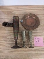 Four metal items on wooden surface: tall brass vase with wood, two brass goblets, copper ashtray.