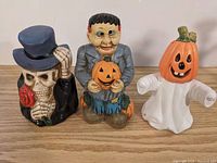Three ceramic Halloween candle holders on a wooden surface, including a skeleton with top hat, Frankenstein figure holding a pumpkin, and ghost with pumpkin head.