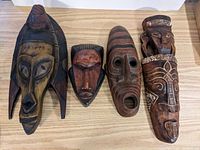 Five carved wooden masks arranged horizontally on a light wood surface, showing different facial details and styles.