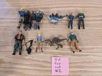 Lot of 8 Jurassic Park action figures, 2 dinosaur toys, and several accessories laid out on a wooden surface marked with a note 'JP Toy Lot #2'.