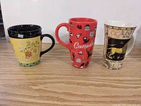 Three large ceramic mugs placed side by side on a wooden surface: Indigo dog mug yellow and black, red Canada-themed mug, and cream Leo zodiac mug.