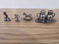Wide shot of four pewter items: three small figurines (boy with watering can, child in motion, rocking horse) and salt and pepper shakers with tray on a wooden surface.