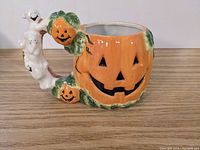 Front-side view of an orange ceramic pumpkin mug with carved facial features and ghosts on the handle
