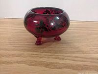 Side view of the ruby red Viking glass bowl with black floral design on its round body and three feet.