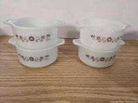 Four small white milk glass casseroles with brown floral pattern stacked in two sets of two, showing front and side views with handles visible.
