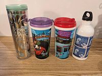 Front view of four drink containers: clear Mickey Mouse and Pluto tumbler, purple lid Disney tumbler, red lid Disneyland-themed tumbler, white Snoopy water bottle